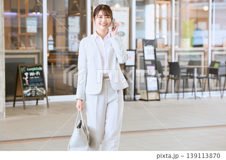 スマートフォンで通話しながら歩く白スーツ姿の若い女性ビジネスパーソン・営業職の外回りイメージ 139113870