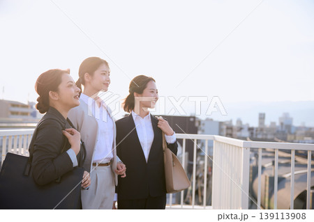 歩道橋の上で前向きに微笑む3人の若い女性ビジネスパーソン・スーツ姿の就活生が朝日を浴びて未来を見つめ 歩道橋の上で前向きに微笑む3人の若い女性ビジネスパーソン・スーツ姿の就活生が朝日を浴びて未来を見つめ 139113908
