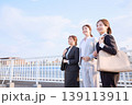橋の上で前向きに微笑む3人の若手女性ビジネスパーソン・スーツ姿で通勤する会社員 139113911