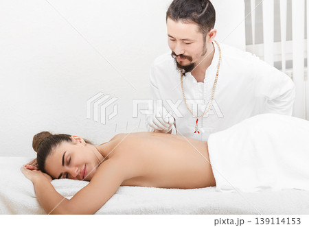 Reflexologist asian race doing acupuncture in the spine of a beautiful woman in a spa salon. Treatment of pain in the back and spine. Woman relaxes and enjoys acupuncture 139114153