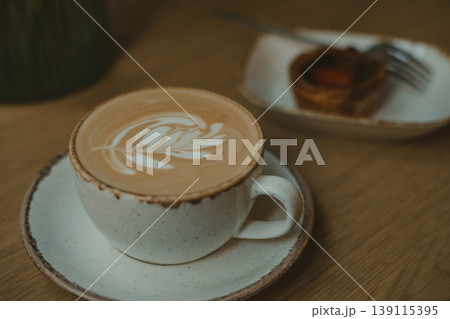Moody high angle shot of latte art in ceramic cup and small tart on wooden table. Aesthetic cafe morning, authentic slow living, rustic breakfast routine, cozy atmospheric cafe lifestyle interior. 139115395