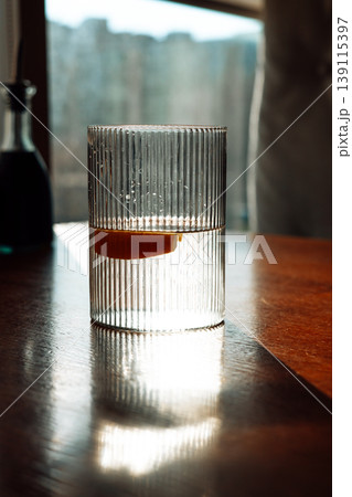 Cinematic close-up of fluted water glass with lemon slice on wooden table. Soft morning sunlight, shadow play, minimalist aesthetic, elegant glassware 139115397