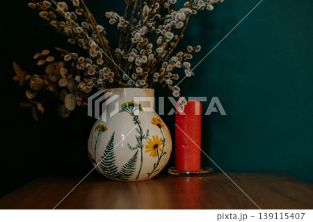 Spring interior still life with pussy willow branches in floral ceramic vase and red candle. Moody aesthetic, farmhouse decor, artisanal pottery, easter atmosphere, cozy home styling on dark teal. Spring interior still life with pussy willow branches in floral ceramic vase and red candle. Moody aesthetic, farmhouse decor, artisanal pottery, easter atmosphere, cozy home styling on dark teal. 139115407
