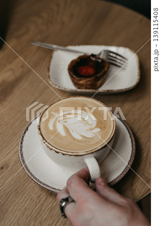 Close up of person hand holding cup of cappuccino with latte art and pastel de nata Portugal dessert on wooden table. Cafe morning, breakfast Close up of person hand holding cup of cappuccino with latte art and pastel de nata Portugal dessert on wooden table. Cafe morning, breakfast 139115408