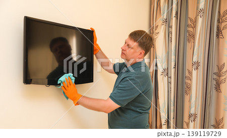 A man carefully wipes dust from a TV screen while cleaning his house 139119729