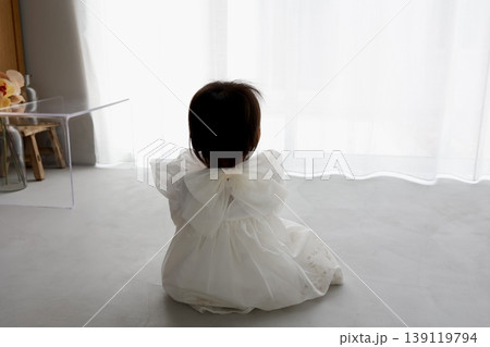 白いドレスの1歳児の後ろ姿。白いカーテンと明るい室内 白いドレスの1歳児の後ろ姿。白いカーテンと明るい室内 139119794