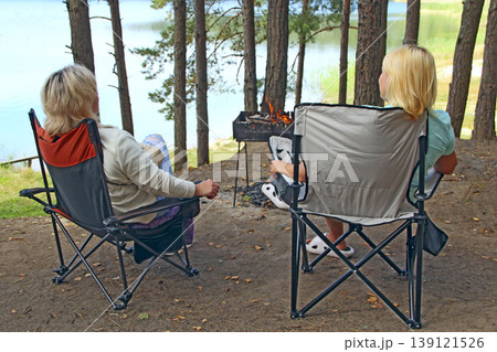 Two women relaxing at picnic near forest lake. Women have a rest by fire Two women relaxing at picnic near forest lake. Women have a rest by fire 139121526