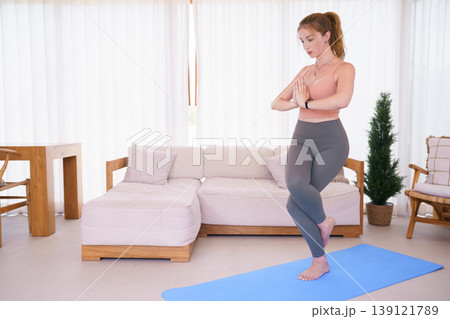 Young woman practicing eagle pose yoga on mat in living room Young woman practicing eagle pose yoga on mat in living room 139121789