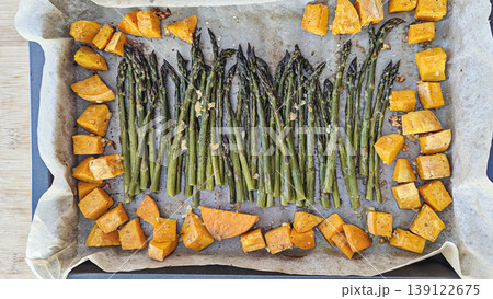 Overhead flat lay of roasted asparagus spears and sweet potato cubes arranged on metal baking sheet, warm tones, rustic healthy meal 139122675