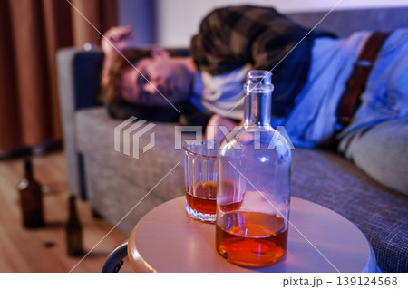 Bottle and glass of alcohol on a table in the foreground. In the blurred background, an unconscious drunk man is asleep on a sofa. Concept of alcoholism, addiction, and crisis. Bottle and glass of alcohol on a table in the foreground. In the blurred background, an unconscious drunk man is asleep on a sofa. Concept of alcoholism, addiction, and crisis. 139124568