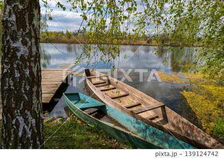 Two wooden boats are nestled by the calm lake, surrounded by lush greenery and reflecting clouds in the serene water Two wooden boats are nestled by the calm lake, surrounded by lush greenery and reflecting clouds in the serene water 139124742