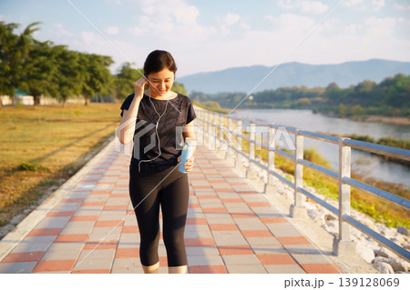 Healthy Asian woman wearing earphones walking exercise on riverside path in morning sunlight 139128069