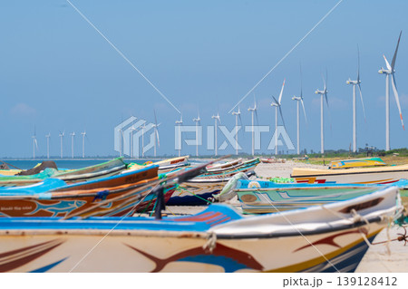 Colorful fishing boats and offshore wind turbines under a clear blue sky. Coastal wind farm on Talaimannar beach, Sri Lanka. Colorful fishing boats and offshore wind turbines under a clear blue sky. Coastal wind farm on Talaimannar beach, Sri Lanka. 139128412