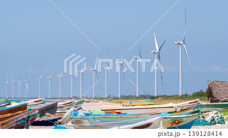 Colorful fishing boats and offshore wind turbines under a clear blue sky. Coastal wind farm on Talaimannar beach, Sri Lanka. Colorful fishing boats and offshore wind turbines under a clear blue sky. Coastal wind farm on Talaimannar beach, Sri Lanka. 139128414