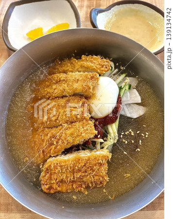 Cold naengmyeon noodles served in a chilled broth with sliced vegetables and egg, paired with crispy golden donkatsu, creating a balance of refreshing and savory flavors in one meal 139134742