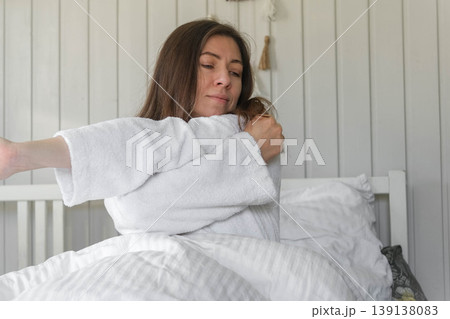 Woman stretching in bed, morning light, soft robe, relaxed expression, cozy white bedding, calm bedroom, waking up before remote work, natural light from the window, quiet moment of self-care. Banner 139138083