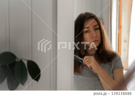 A woman combs her hair in front of a mirror. She's tidying herself up. She's wearing a casual gray T-shirt. Her expression is focused, as she prepares for the workday. Self-care. Space for text. A woman combs her hair in front of a mirror. She's tidying herself up. She's wearing a casual gray T-shirt. Her expression is focused, as she prepares for the workday. Self-care. Space for text. 139138086