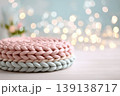 Knitted cushion podium on table, spring blossom bokeh background, pastel tones. Wool pad on surface. Soft pedestal. Cozy stage for promotional imagery. Showcase display case. Front view. Mockup ad 139138717
