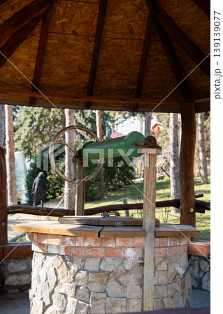 Old water well with metal handle and wooden structure under a gazebo roof in a park surrounded by trees in daylight. Rustic equipment, outdoor architecture, history Old water well with metal handle and wooden structure under a gazebo roof in a park surrounded by trees in daylight. Rustic equipment, outdoor architecture, history 139139077