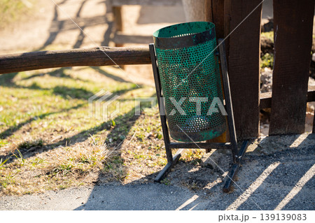 Green metal trash bin standing near wooden fence in a zoo park area with grass and sunlight casting shadows on ground. Cleanliness, environment, waste management Green metal trash bin standing near wooden fence in a zoo park area with grass and sunlight casting shadows on ground. Cleanliness, environment, waste management 139139083