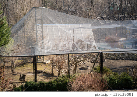 Large animal enclosure covered with protective netting in a zoo with trees, fences and dry ground under daylight. Wildlife habitat, captivity, environment 139139091
