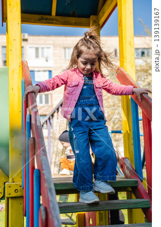 Little girl in pink jacket walking down colorful playground stairs and holding rails outdoors. Concept of childhood activity and playground safety 139139167