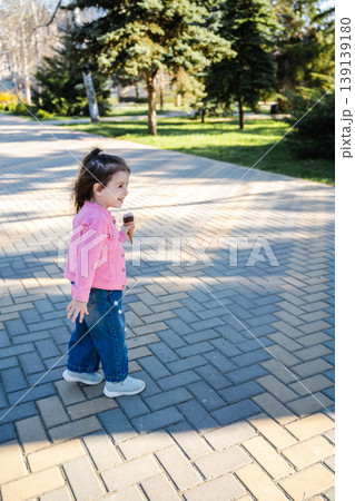 Little girl in pink jacket walking on paved path in park while holding ice cream cone. Concept of childhood walk and outdoor leisure Little girl in pink jacket walking on paved path in park while holding ice cream cone. Concept of childhood walk and outdoor leisure 139139180