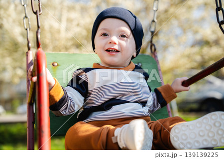 Little boy in hat sitting on swing seat holding bars and smiling in sunny playground environment. Concept of childhood joy and outdoor activity 139139225