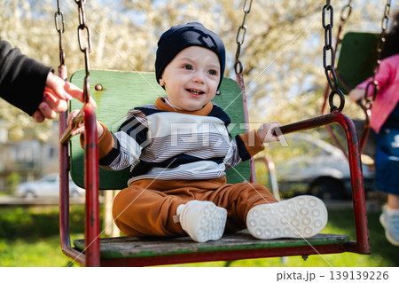 Little boy in hat sitting on swing seat holding bars while adult hand pushes in outdoor playground. Concept of childhood play and parental support 139139226