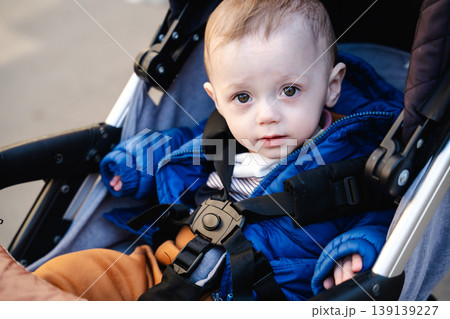 Baby sitting in stroller wearing blue jacket and safety belt looking at camera outdoors. Concept of childcare and safety 139139227