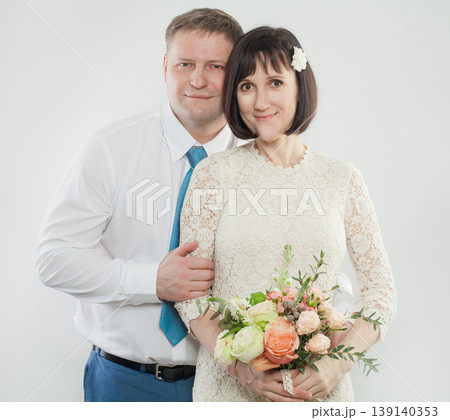 A woman holds a bouquet while hugging a man from behind in a studio. Both smile and look happy. A woman holds a bouquet while hugging a man from behind in a studio. Both smile and look happy. 139140353