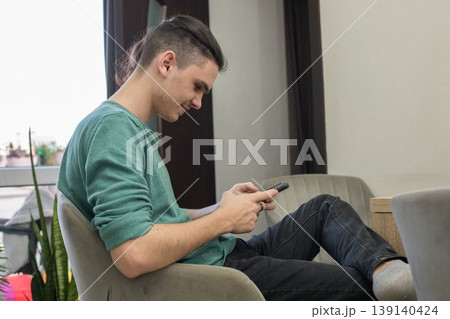 Young man sits in chair and looks at phone in modern indoor space during daytime Young man sits in chair and looks at phone in modern indoor space during daytime 139140424