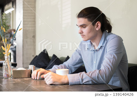 A man is seated at a table in a cafe. He holds a cup and appears to be lost in thought. A man is seated at a table in a cafe. He holds a cup and appears to be lost in thought. 139140426