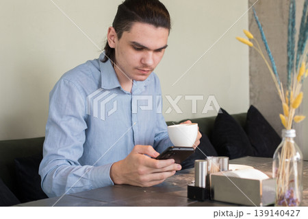 A young man is sitting at a table in a cafe. He looks at his phone and holds a cup A young man is sitting at a table in a cafe. He looks at his phone and holds a cup 139140427