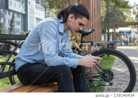 A young man sits on a bench in a park during the day.  139140434