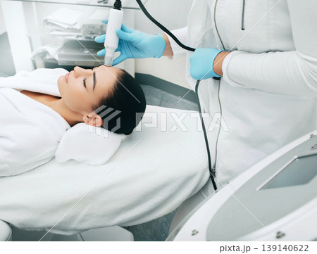 Beautician using machine no-needles mesotherapy smoothes wrinkles of the skin of the female forehead . Painless skin rejuvenation method Beautician using machine no-needles mesotherapy smoothes wrinkles of the skin of the female forehead . Painless skin rejuvenation method 139140622