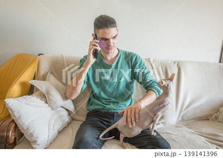 A young man sits on a couch holding his phone. A cat sits beside him on the couch A young man sits on a couch holding his phone. A cat sits beside him on the couch 139141396