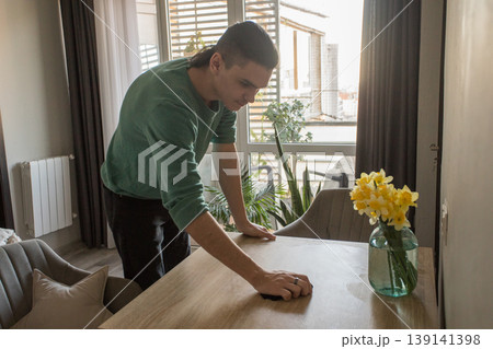 A man is wiping the surface of a wooden table in a well-lit room. A man is wiping the surface of a wooden table in a well-lit room. 139141398