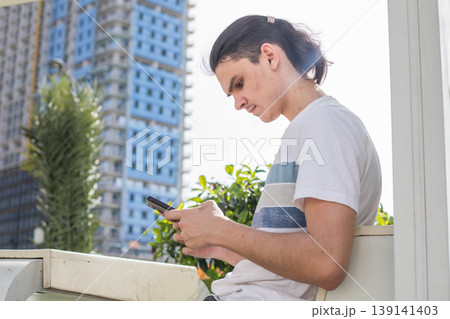 A young man is seated on a bench, looking at his phone with focus. 139141403