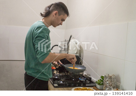 Young man cooking a dish in a kitchen while preparing a meal with fresh ingredients and tools 139141410