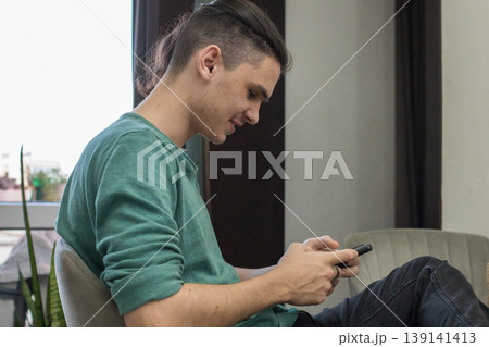 A young man is sitting in a chair and looking at his phone in a well-lit indoor area. 139141413