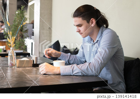 A young man is sitting at a table in a cafe. He looks at his phone and holds a cup A young man is sitting at a table in a cafe. He looks at his phone and holds a cup 139141418