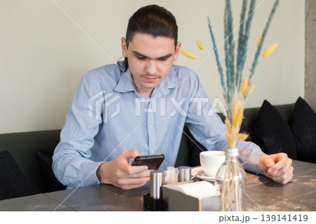 A young man is sitting at a table in a cafe. He looks at his phone and holds a cup A young man is sitting at a table in a cafe. He looks at his phone and holds a cup 139141419