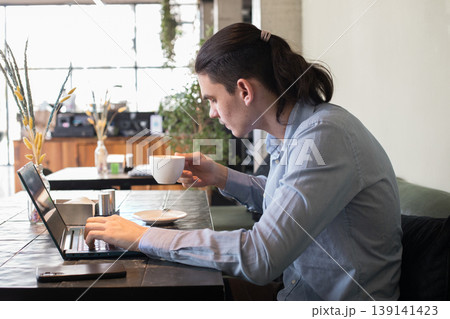 A young man sits at a cafe table focused on his laptop while sipping coffee. 139141423
