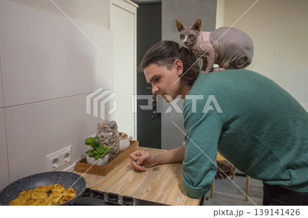 A man stands in a kitchen near a stove while a cat sits on his shoulder. A man stands in a kitchen near a stove while a cat sits on his shoulder. 139141426