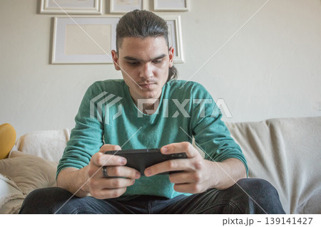 Teenager plays video game on mobile phone while sitting on couch in living room in daylight Teenager plays video game on mobile phone while sitting on couch in living room in daylight 139141427