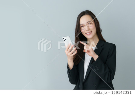 グレーバックの眼鏡をかけたスマホを持つ若いスーツの日本人女性 グレーバックの眼鏡をかけたスマホを持つ若いスーツの日本人女性 139142351