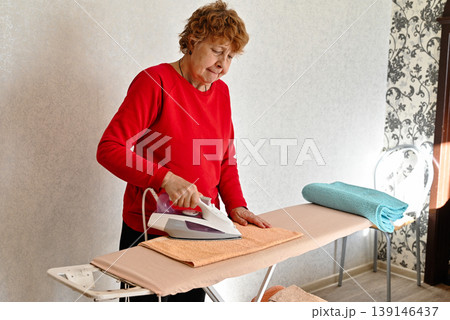 Middle age grey-haired woman talking on smartphone ironing clothes at laundry room Middle age grey-haired woman talking on smartphone ironing clothes at laundry room 139146437