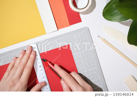 Hands using utility knife and ruler to cut red paper on cutting mat. Workspace for DIY paper crafts. Hands using utility knife and ruler to cut red paper on cutting mat. Workspace for DIY paper crafts. 139146814