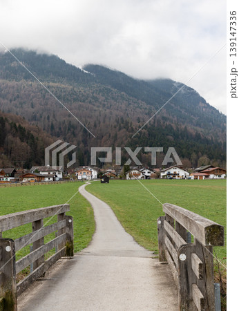 Rural path leading through green meadow toward alpine village with mountains and cloudy sky in Bavaria Germany Vertical Rural path leading through green meadow toward alpine village with mountains and cloudy sky in Bavaria Germany Vertical 139147336